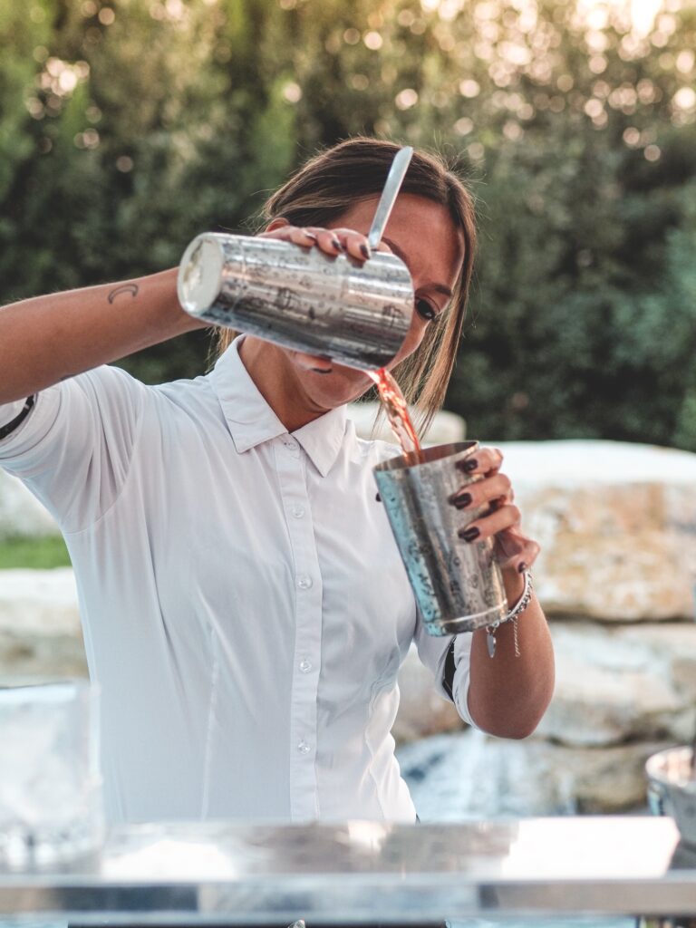 blonde female bartedenr staff make red cocktail drink on decorated low glass outdoors in event