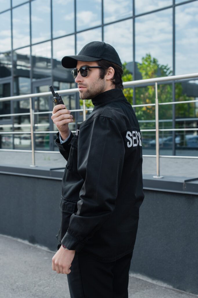 young security man in uniform, cap and sunglasses talking on walkie-talkie near building